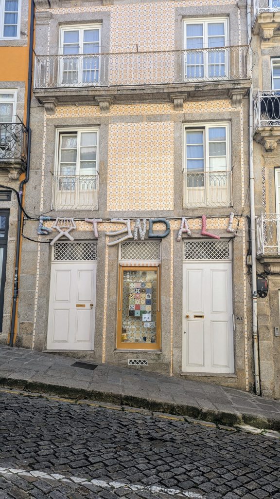 A three-story building features a tiled facade with two shuttered windows on each upper floor and two white doors on the ground floor. A sign with colorful letters hangs above the doors.