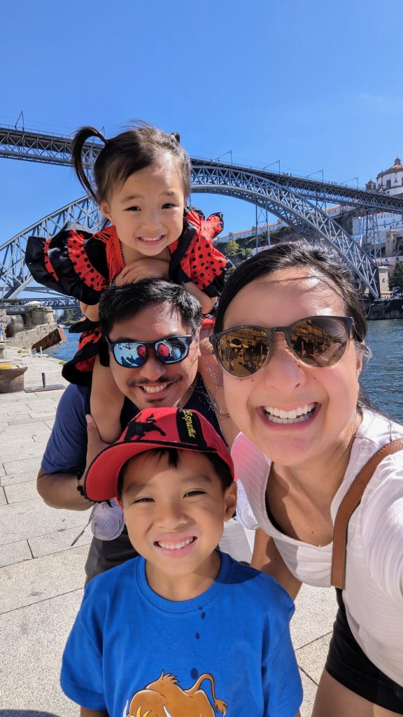 A family of four, including a man, woman, and two children, smiles for a selfie by a river with an arched metal bridge in the background on a sunny day.