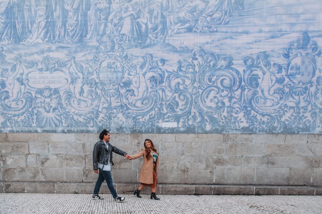 A man and a woman hold hands and walk past a large, church wall covered in blue and white azulejo tiles.