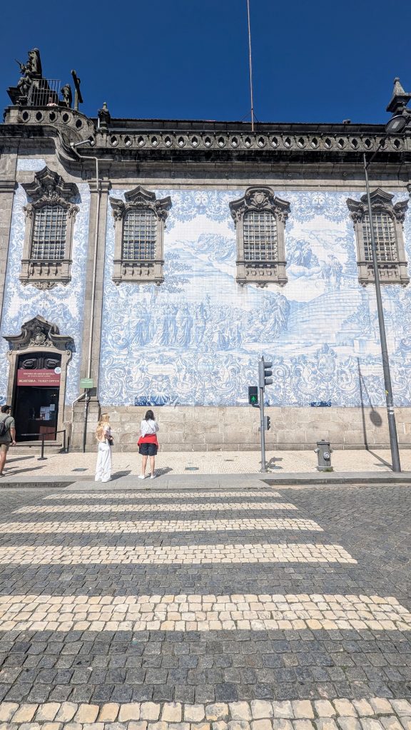 Two people walk on a cobblestone crosswalk in front of a historic building featuring an extensive blue and white tile mural under a clear blue sky.