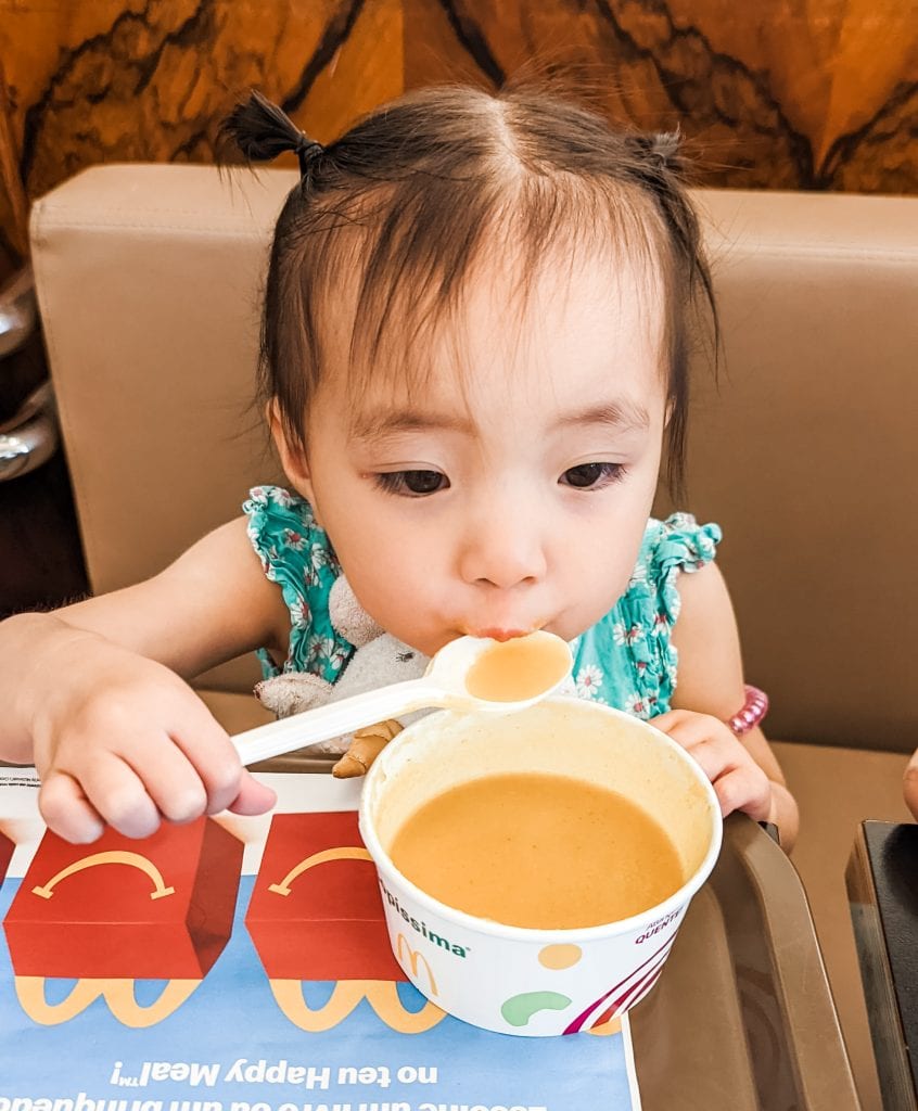 A toddler with pigtails sits at a table, eating soup from a white container using a spoon.