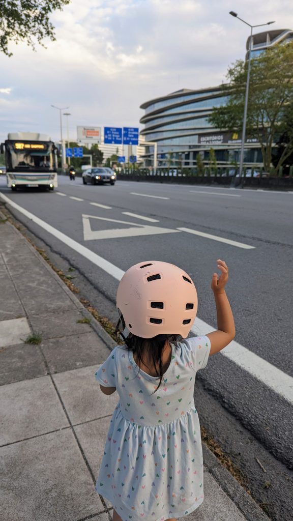 A child wearing a light pink helmet and a dress flags a bus down from the sidewalk beside a multi-lane road.