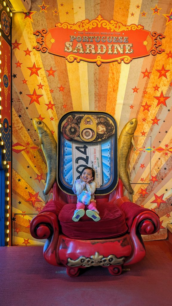 A child sits on a large, ornate red chair in front of a vibrant backdrop featuring fish and stars, with a sign that reads "The Fantastic World of the Portuguese Sardine"