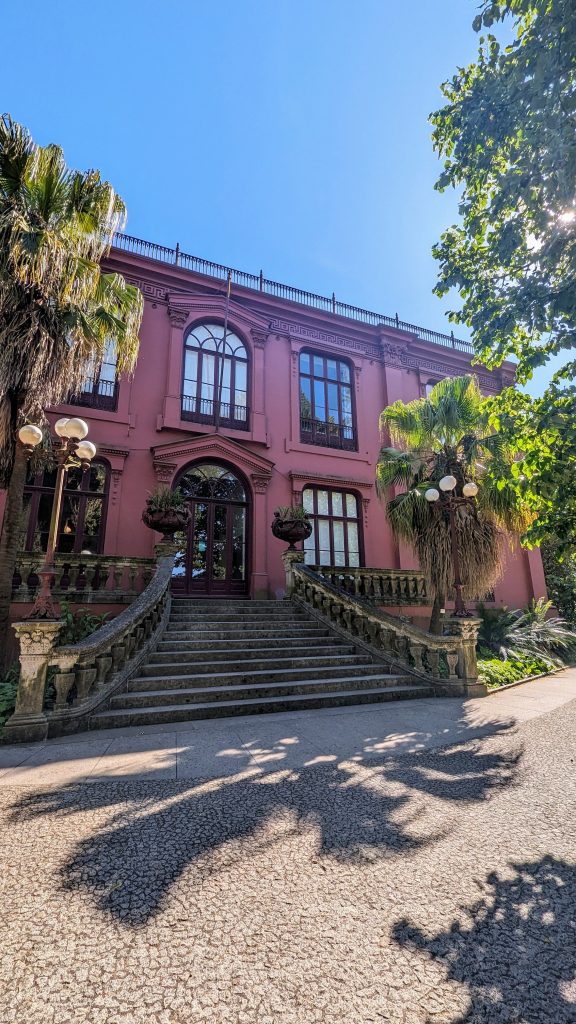 A large pink building with arched windows and a grand staircase, surrounded by trees and palm plants in a well-lit, sunny setting.