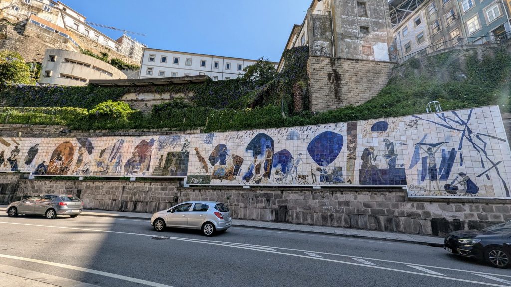Cars drive along a street beside a long tiled mural depicting various scenes, while historic buildings and lush greenery rise in the background.