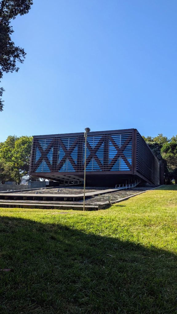 A modern building with a geometric design featuring blue panels and dark framing sits on a lush, grassy area with trees in the background under a clear blue sky.