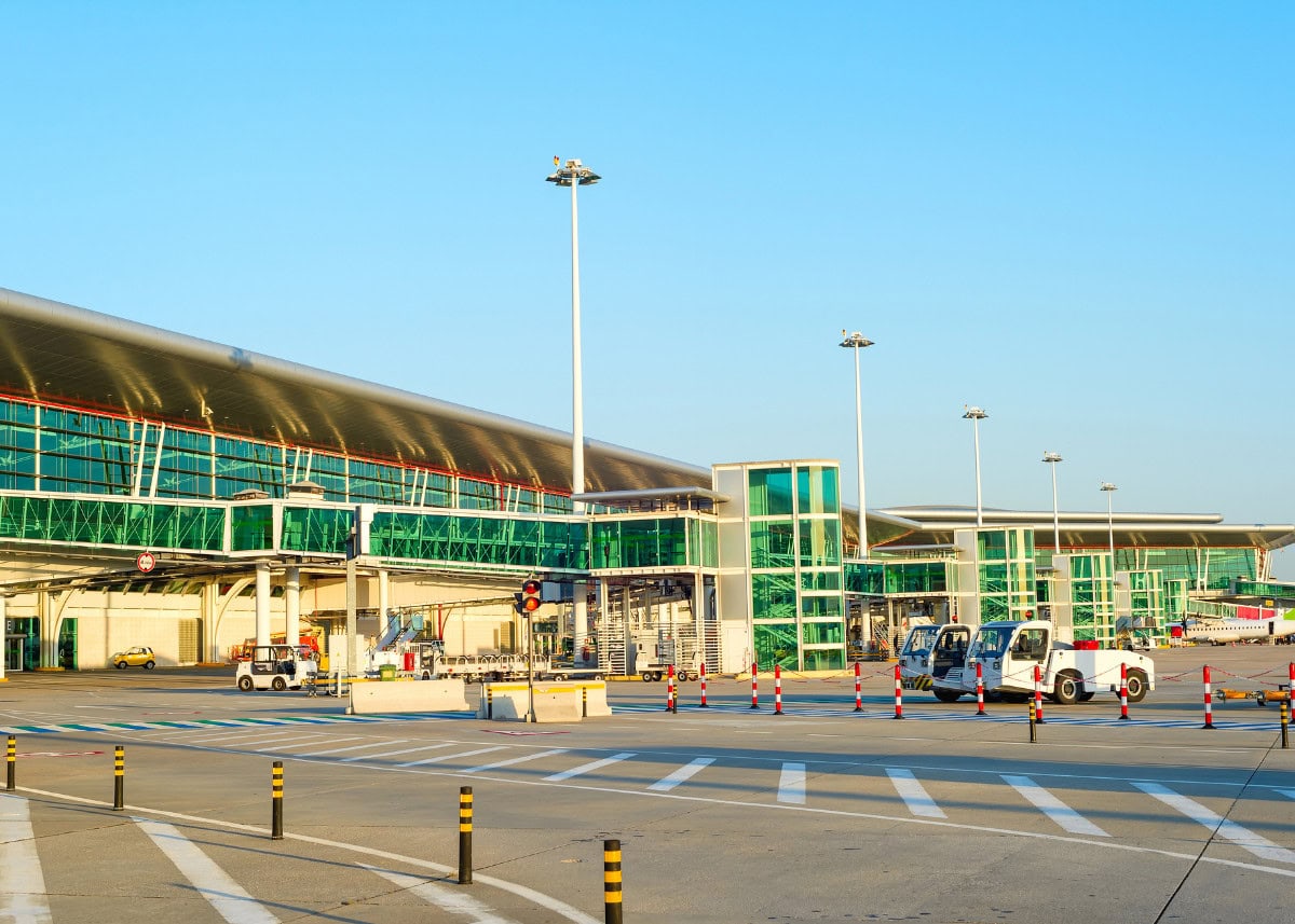 An airport terminal building with several glass facades and modern architecture. Service vehicles are parked on the tarmac, and traffic cones line a section of the area.