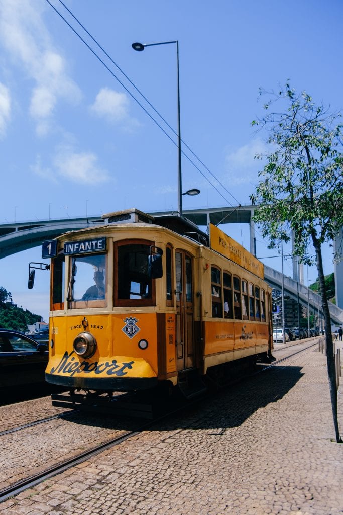 Yellow tram marked "Infante" on Line 1 moves along a cobblestone street near a modern bridge and a tree on a clear day.
