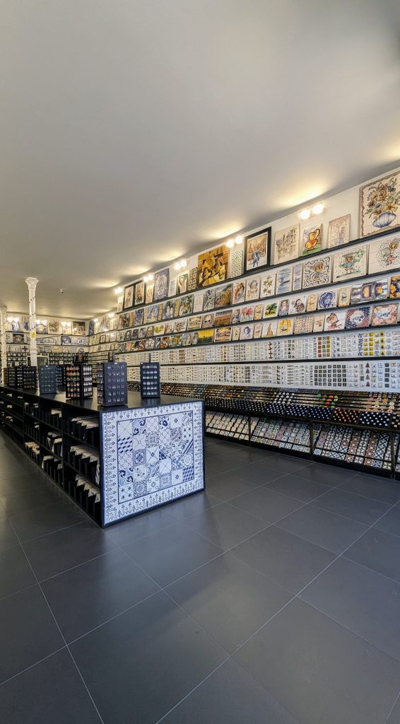 A well-lit store interior with shelves and walls displaying a variety of decorative tiles and artworks. The floor is tiled in gray, and the ceiling has white lighting fixtures.