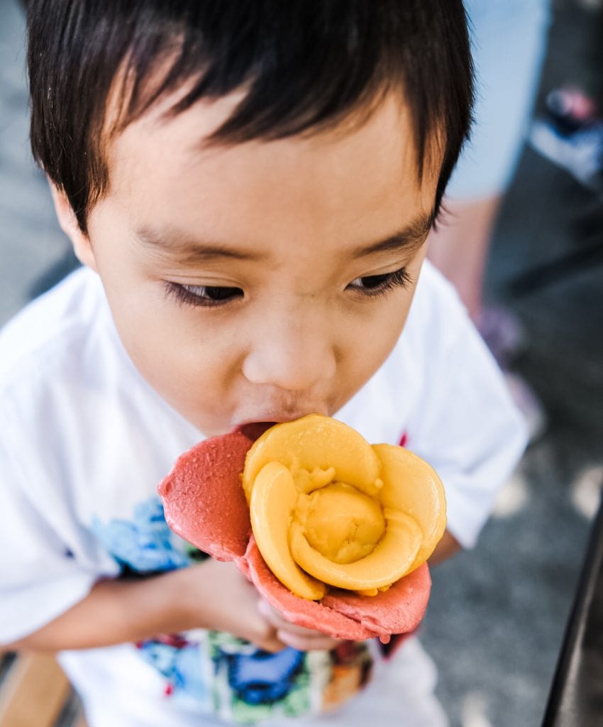 A young child in a white shirt is eating a flower-shaped ice cream cone with yellow and pink scoops.