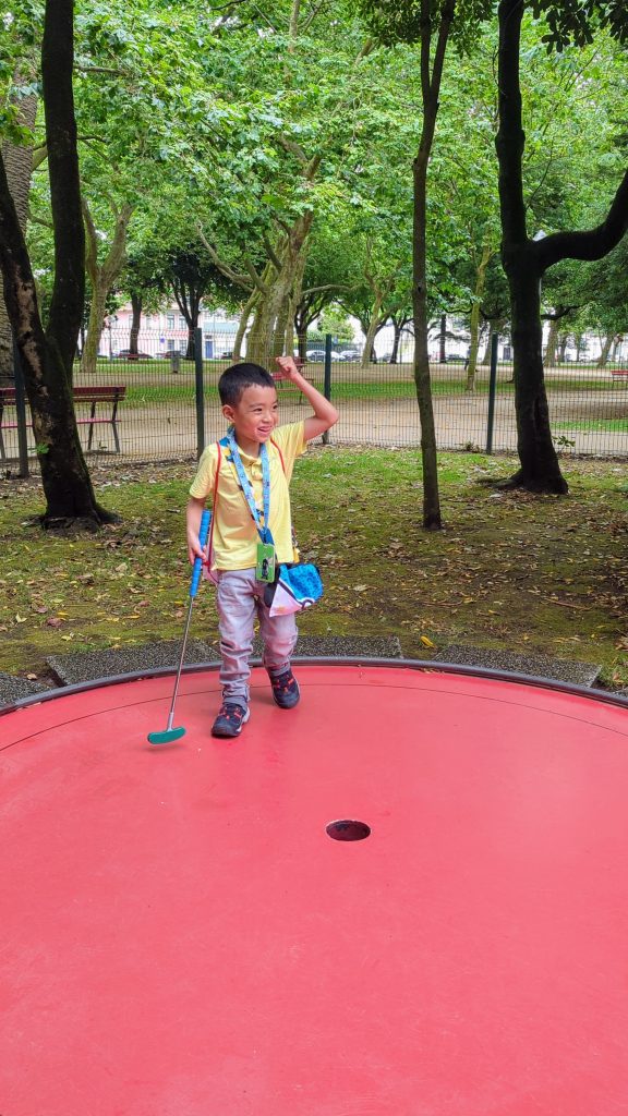 A child stands on a red circular platform, holding a mini golf putter and smiling. Trees and a fence fill the background.