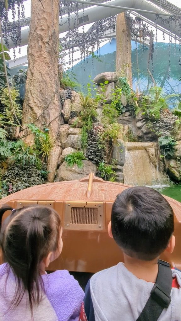 Two children sit in a boat facing a scenic, lush environment with rocks, plants, and a small waterfall.