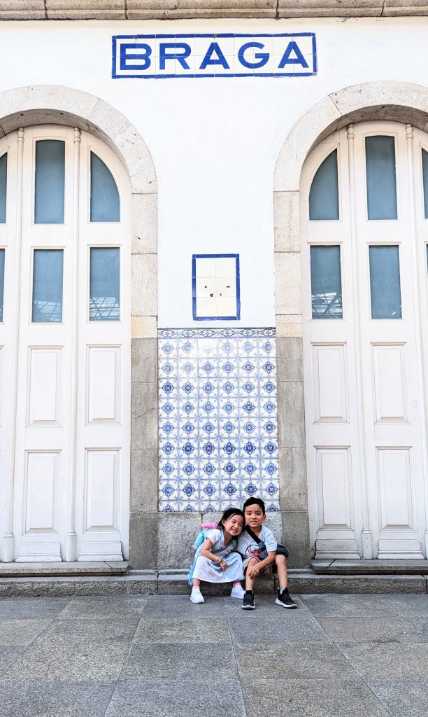 Two children sit on the steps in front of a white building with twin arched doors and a large "BRAGA" sign above them. The section between the doors features decorative blue and white tiles.