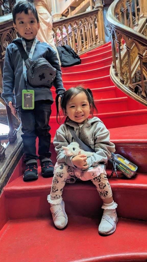 Two children pose on a red-carpeted staircase. The boy stands holding a small bag, and the girl sits on the step below, holding a stuffed toy and smiling. Wooden railings are visible in the background.