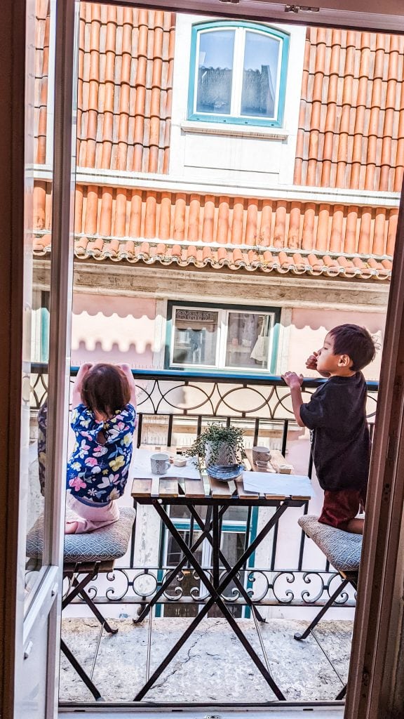 Two young children stand on chairs on a small balcony, looking over the railing. The balcony has a table with potted plants and drinks. There is a tiled roof building with windows in the background.