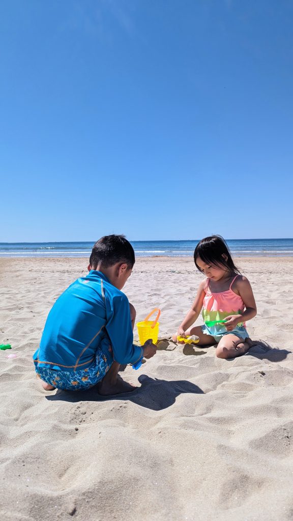 Two children playing with sand toys on a beach under a clear blue sky. The ocean is visible in the background.