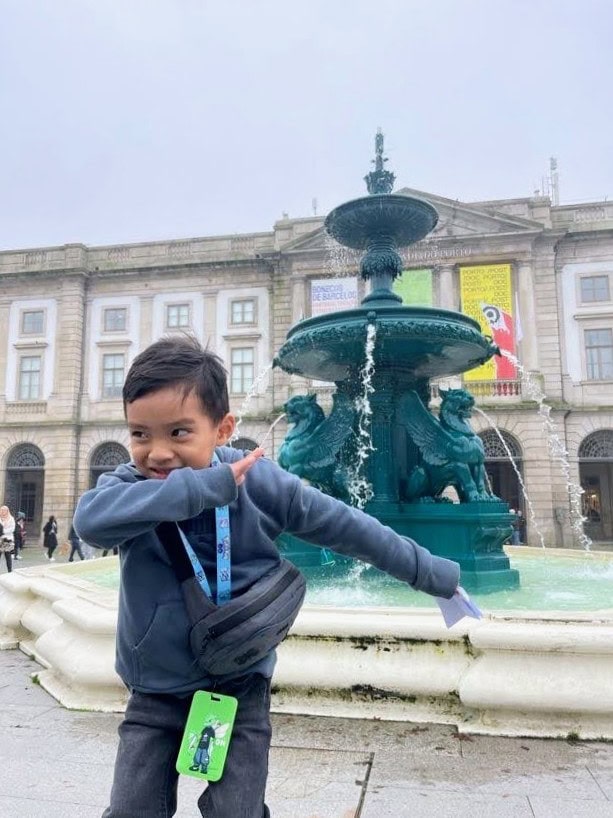 A young boy in a blue sweatshirt performs a dab pose in front of an ornate fountain with building in the background. He wears a crossbody bag and has a green card case hanging around his neck.