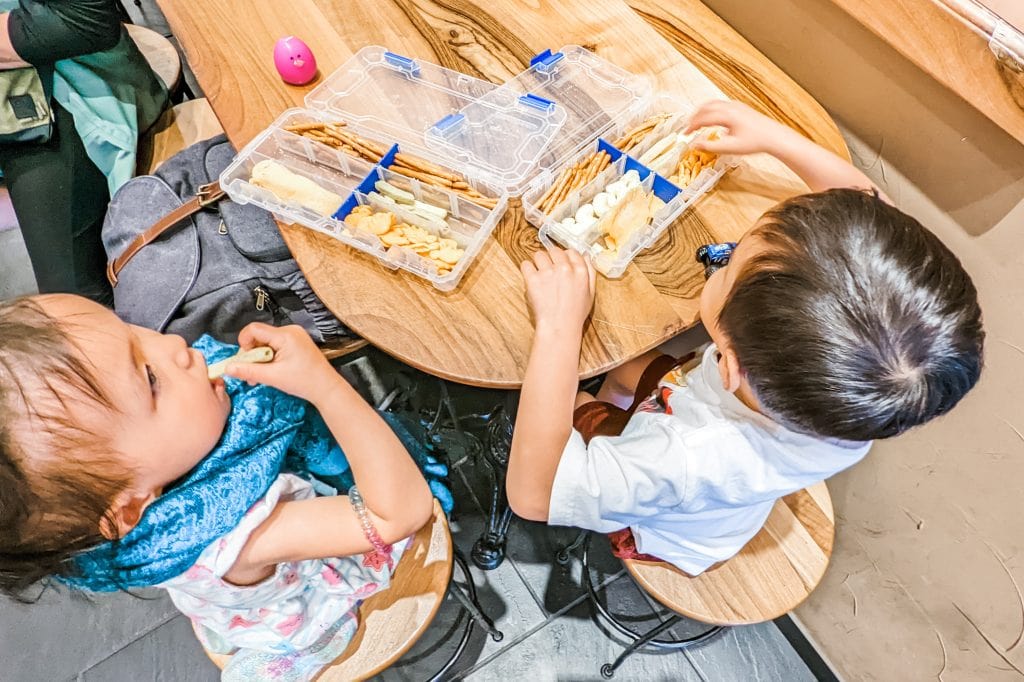 Two children sit at a wooden table with snack boxes containing crackers and cheese. One child is eating while the other looks at the food.