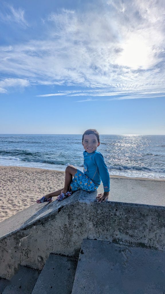 A child in blue swimwear sits on concrete steps overlooking a sunny beach and ocean.
