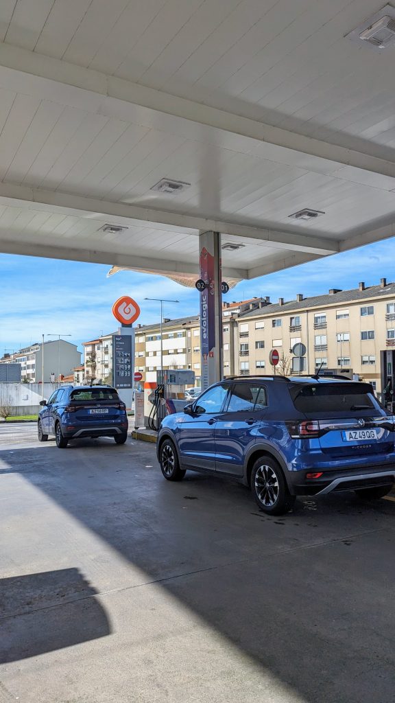 Two blue cars parked at a gas station next to fuel pumps, with buildings in the background under a clear sky.
