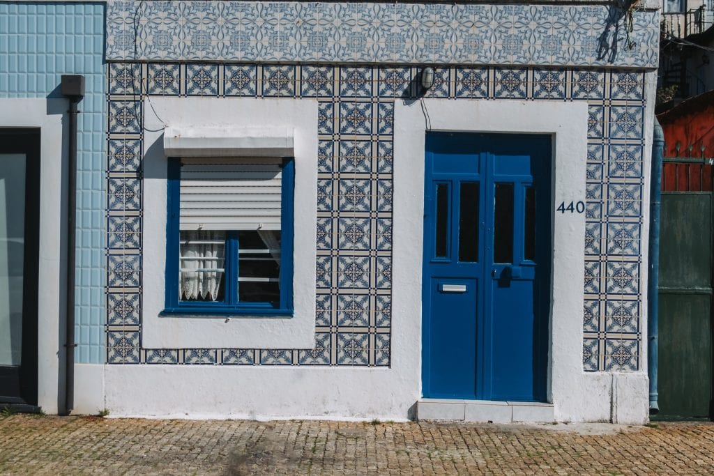A building exterior with a blue and white tiled facade, blue door, blue-framed window with a lowered shutter, and the number 440 beside the door.