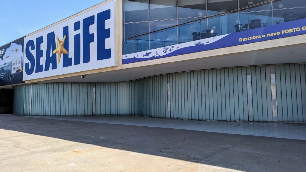 Exterior of a SEA LIFE aquarium featuring the logo on the building. The area below it shows metallic shutters, indicating the entrance. The banner reads "Descubra o novo PORTO".
