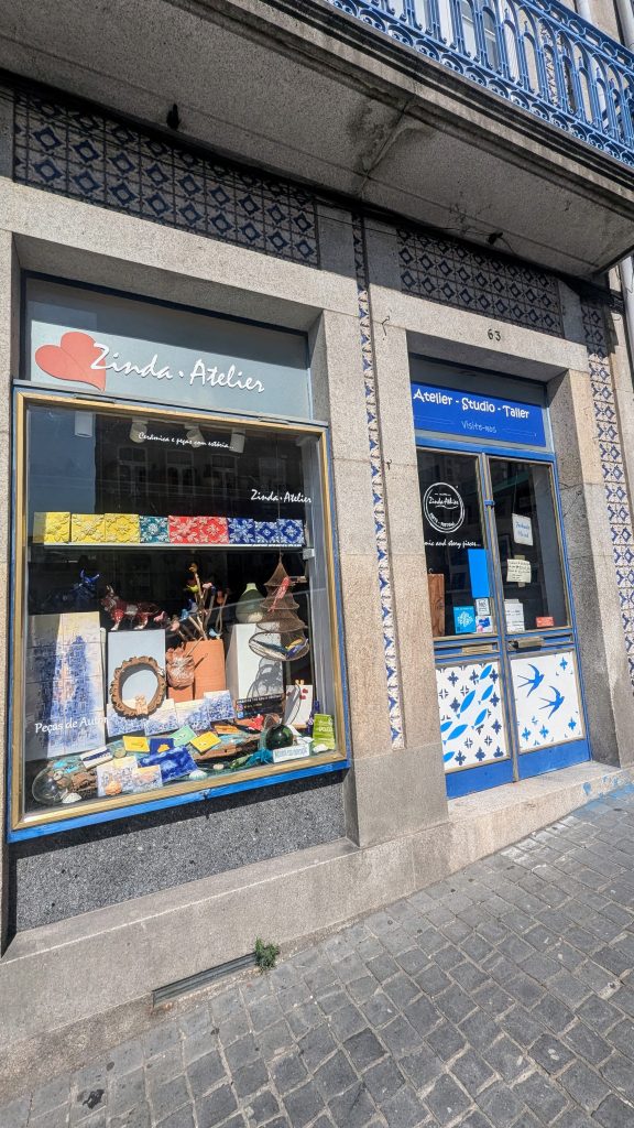 Exterior of "Zinda Atelier" store showcasing a colorful window display of various craft items, located in a building adorned with blue and white tiles. A blue door with glass panels is on the right.