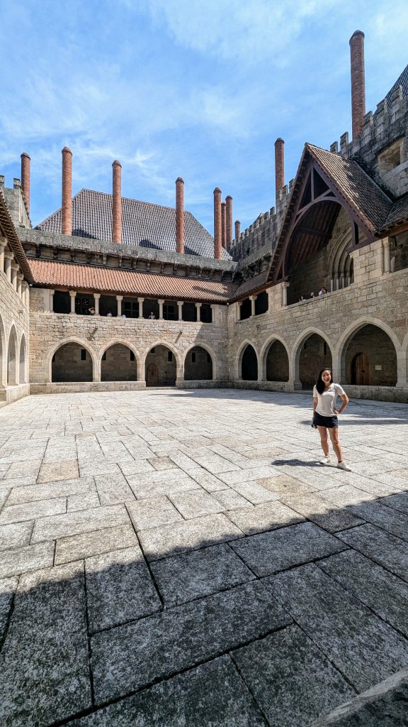 A woman stands in the courtyard of a historic stone building featuring arched doorways and multiple tall chimneys, under a clear blue sky.