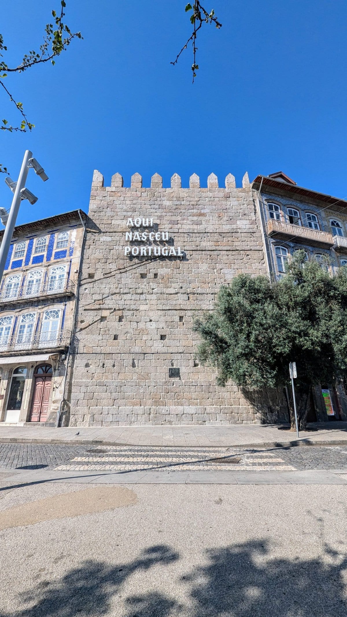 A historic stone building with Portuguese text "AQUI NASCEU PORTUGAL" on its wall. The structure features medieval architectural elements and adjacent modern lampposts, all under a clear blue sky.