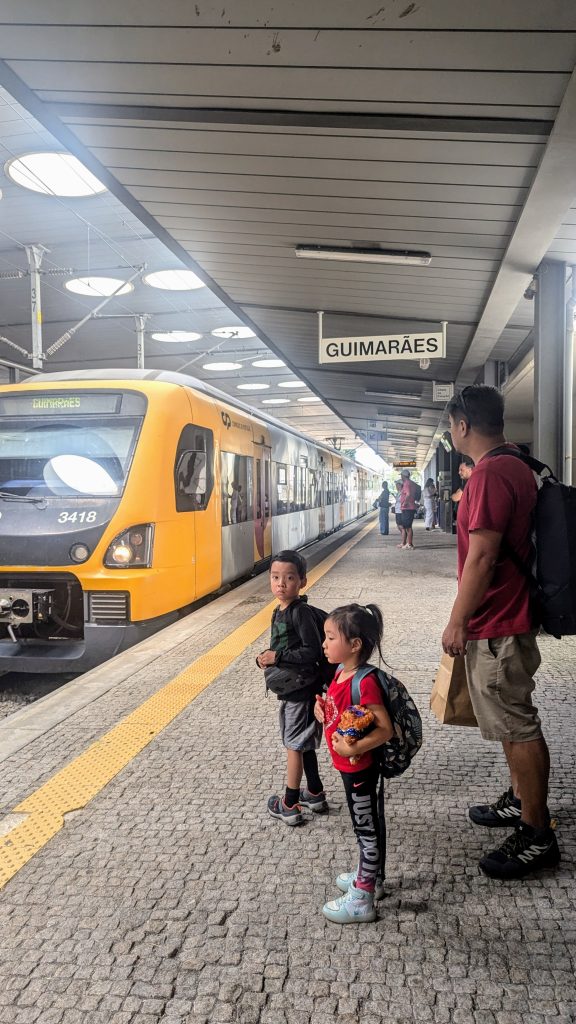 A man and two children stand on a train platform next to a stationary yellow and gray train at Guimarães station. The platform is covered and other passengers are visible in the background.