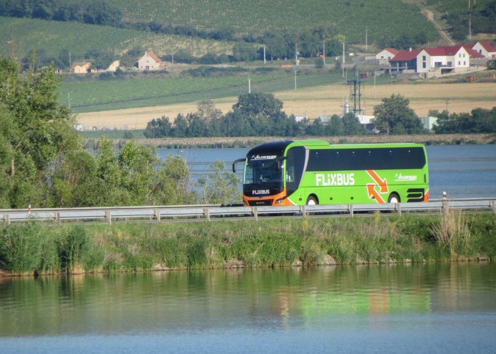 A green and orange FlixBus travels on a road next to a body of water with houses and greenery in the background.