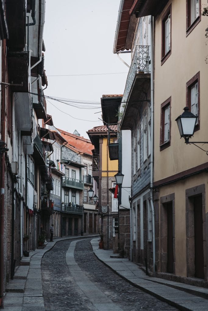 Narrow, empty cobblestone street lined with old buildings featuring balconies and shuttered windows. Overhead, electrical wires crisscross between structures. A single streetlamp is visible.