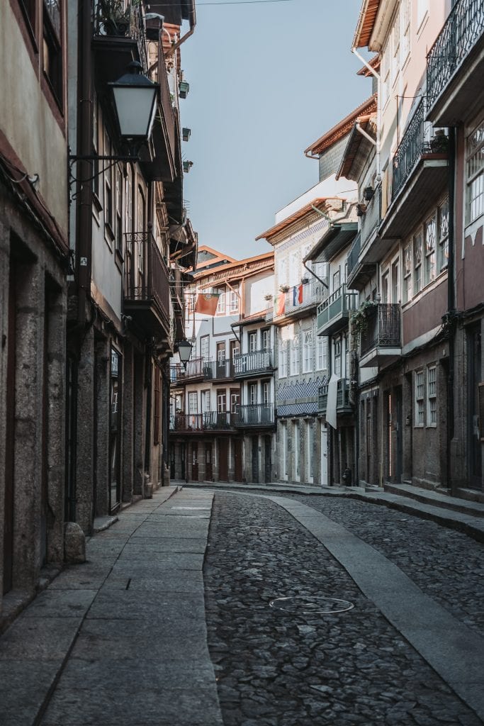 Narrow, empty cobblestone street lined with old, multi-story buildings featuring balconies and overhanging roofs under a blue sky.