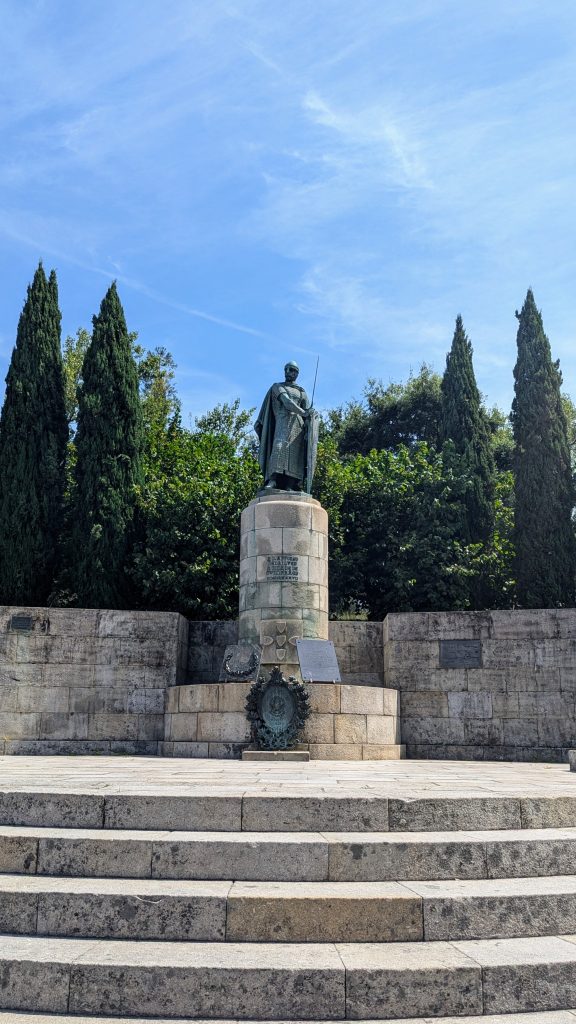 A bronze statue of a historical figure stands atop a stone pedestal surrounded by trees and foliage, with a set of stone steps leading up to it.