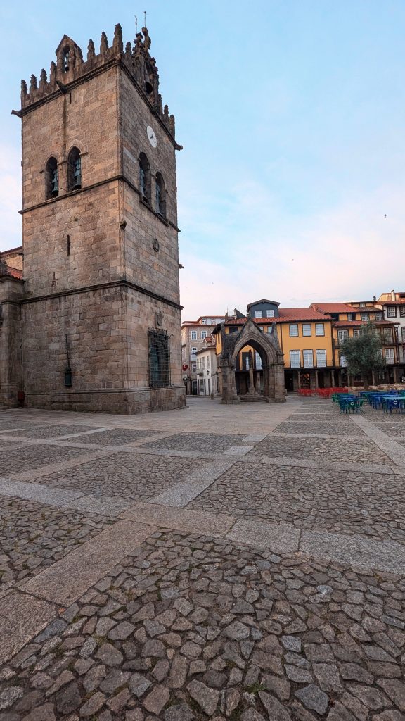A large stone church with a bell tower stands in a cobblestone plaza surrounded by buildings. Blue chairs are set up to the right, and the sky is clear with a soft light.
