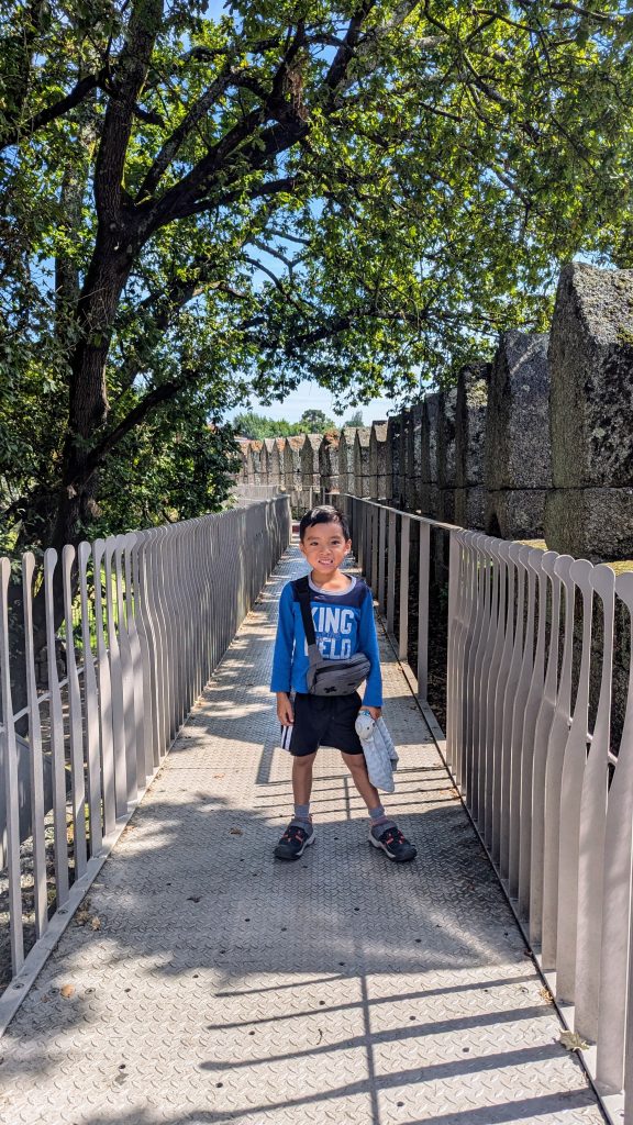 A young boy stands on a narrow metal walkway flanked by railings, with trees and stone structures visible in the background. He wears a blue shirt, dark shorts, and tennis shoes.