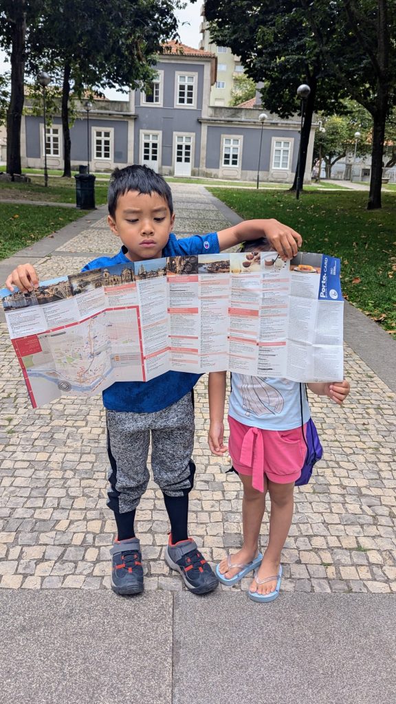 Two children stand on a paved outdoor path, holding a large map that partially covers their faces. Trees and a building in the background.