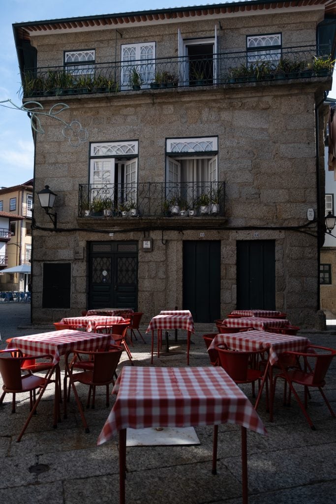 Outdoor café with red and white checkered tablecloths on tables arranged in front of a two-story stone building with balconies and windows. The setting appears to be a quiet street.