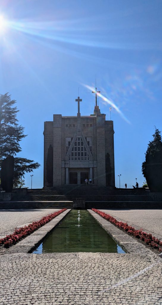 A modern church with a cross and angular facade, viewed with a sunburst sky. A flower-lined water feature leads up to the entrance.