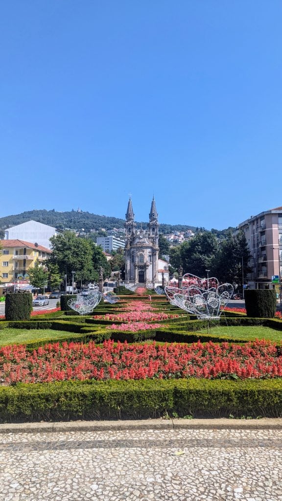 A well-maintained garden with symmetrical flower beds and shaped topiaries, leading up to a church with dual steeples, set against a backdrop of trees on a clear day.