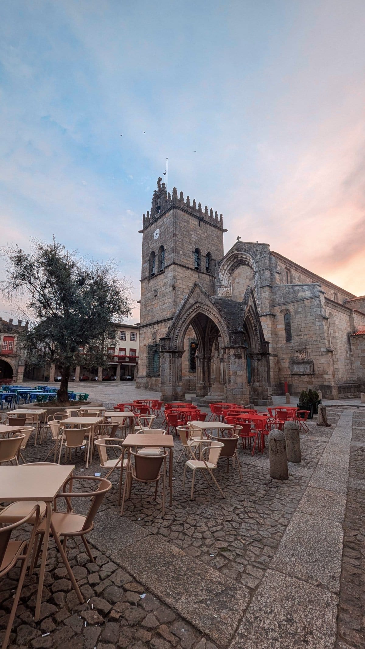 A stone-paved courtyard with empty tables and chairs faces a historic stone church with Gothic architecture under a sunset sky.