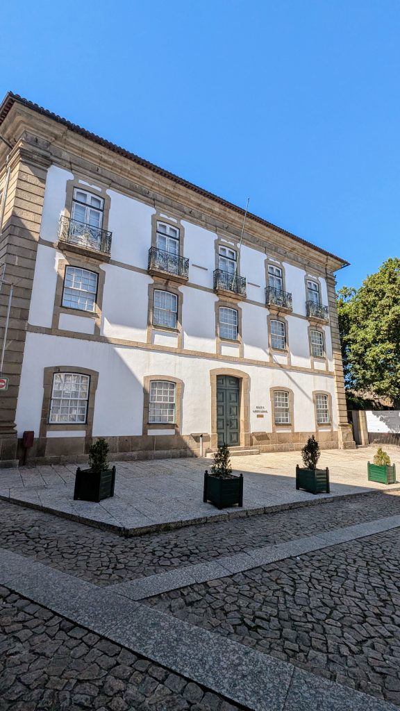 A three-story historical building with white walls, rectangular windows, and a large green door. The building is surrounded by a cobblestone courtyard and potted plants.