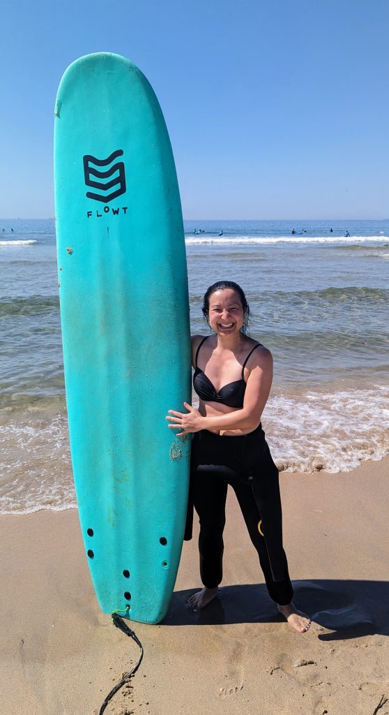 A person in a black swimsuit and wetsuit pants stands on a sandy beach holding a turquoise surfboard, with the ocean and other surfers in the background.