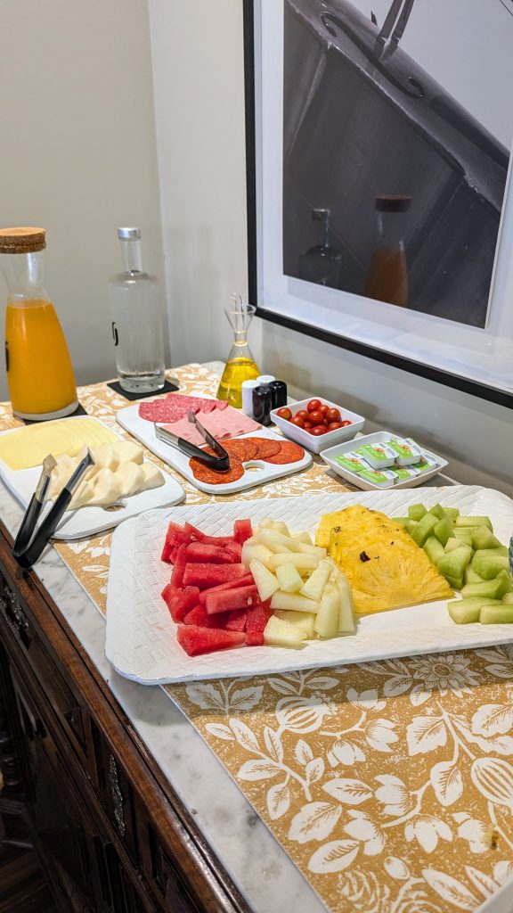 A buffet table with cut fruit, sliced cheese, cold cuts, cherry tomatoes, butter, bottled juice, a glass bottle, a jar, and condiments.