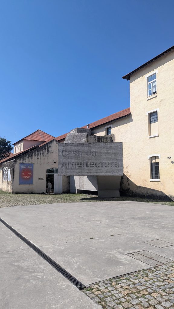 A concrete structure labeled "Casa da Arquitectura" extends from a beige building with a red-tiled roof, under a clear blue sky.