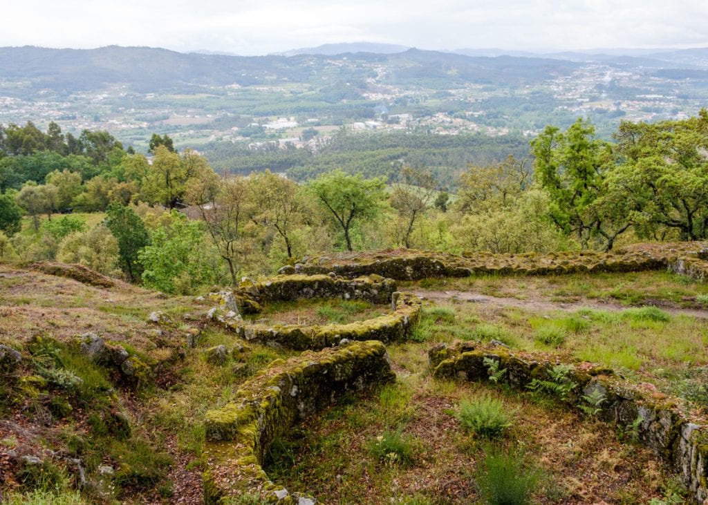 Stone ruins of an ancient settlement on a grassy hill, surrounded by trees, with a vast valley and distant mountains visible in the background.