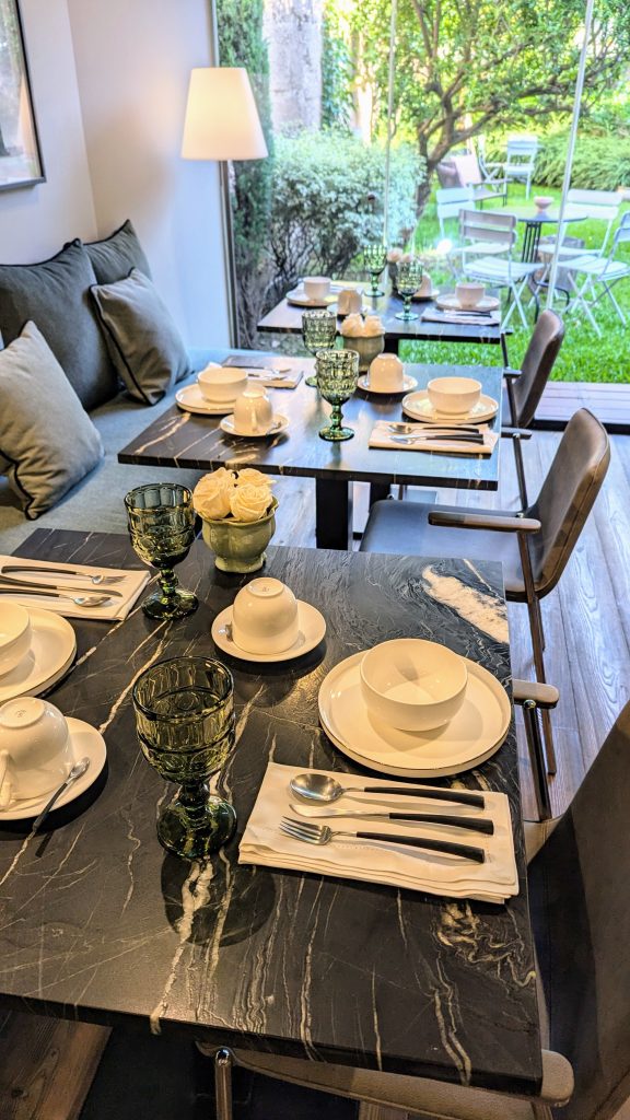 A dining table set with cups, plates, and green glasses in a room with a large window overlooking a green garden, with chairs arranged around the table and a sofa in the background.