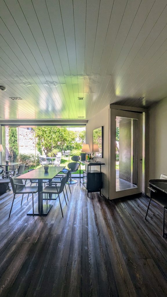 A modern dining room with large windows, a wooden floor, tables and chairs, and a view of a garden outside.