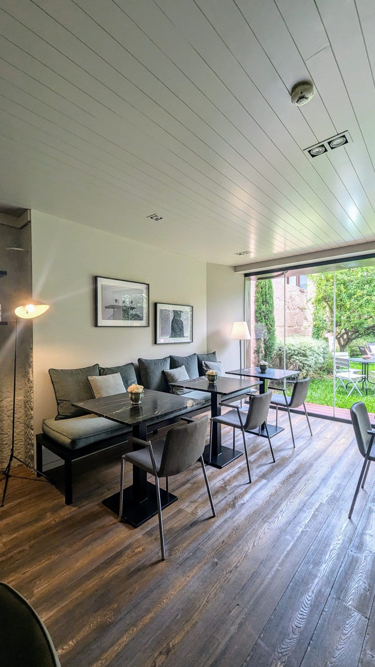 Modern dining area with a large table, cushioned bench seating, chairs, and a floor lamp. Glass doors on the right open to a garden with outdoor seating. Walls display framed artwork.