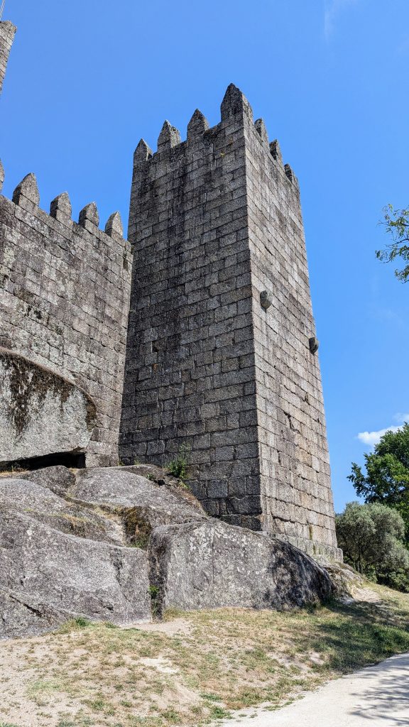 A tall, stone castle tower with crenellations stands under a clear blue sky, adjacent to a rocky hillside and surrounded by grass.